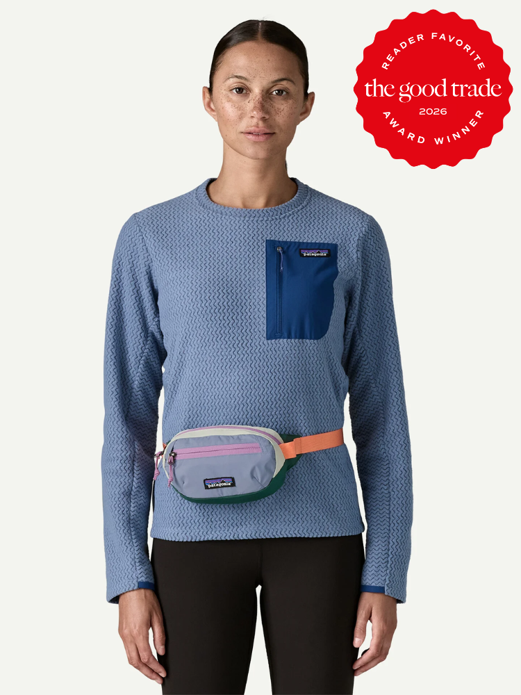 A person wearing a blue textured long-sleeve shirt with a blue chest pocket, black pants, and a pastel-colored fanny pack. A red "The Good Trade Award Winner 2026" badge is in the corner.