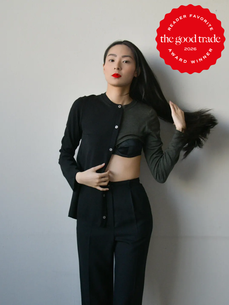 A person wearing a two-tone black top and black trousers stands against a plain wall, adjusting their hair. A "The Good Trade 2026 Award Winner" badge is in the top right corner.