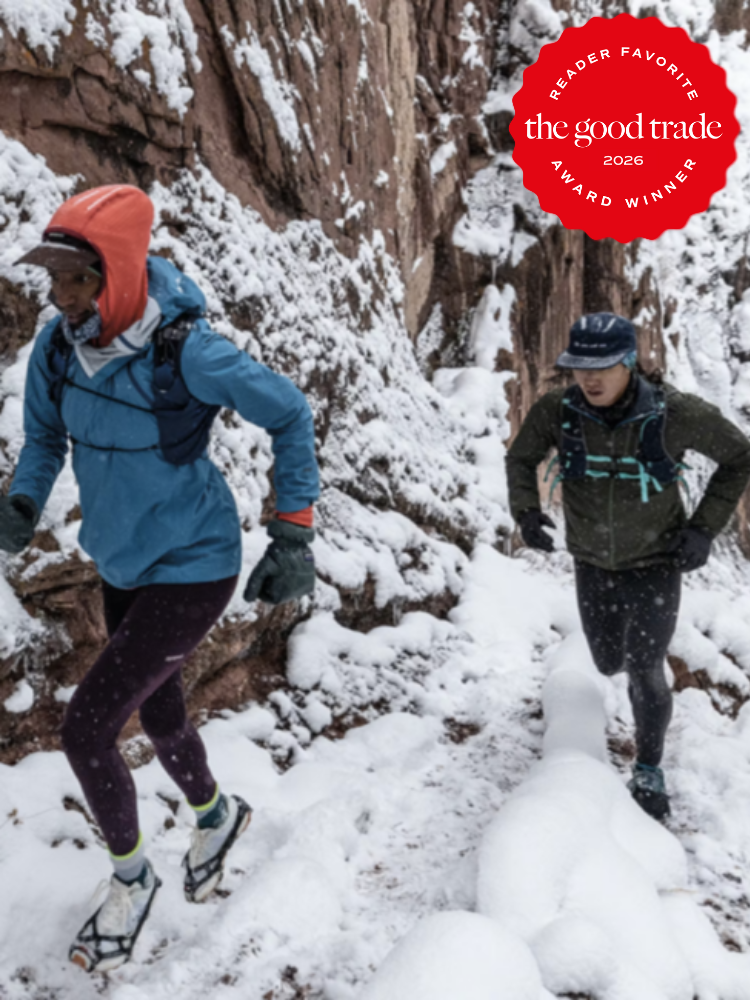 Two people wearing winter gear and backpacks trail run on a snowy, narrow mountain path. A red award badge in the corner reads, "The Good Trade 2026 Reader Favorite Award Winner.
