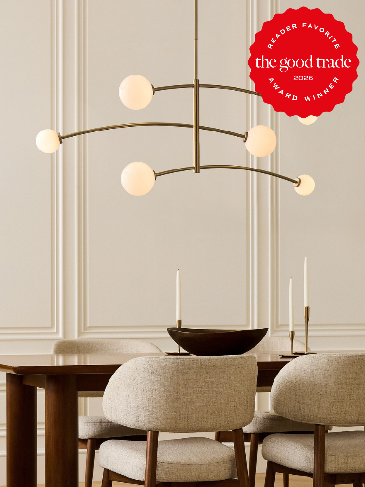Modern dining room with a brass chandelier, beige upholstered chairs, a wooden table set with candlesticks, and an award badge reading “The Good Trade 2026 Reader Favorite.”.