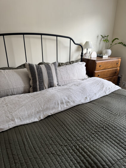 A neatly made bed with green bedding and multiple pillows is pictured alongside a wooden bedside table adorned with a small lamp and plant.