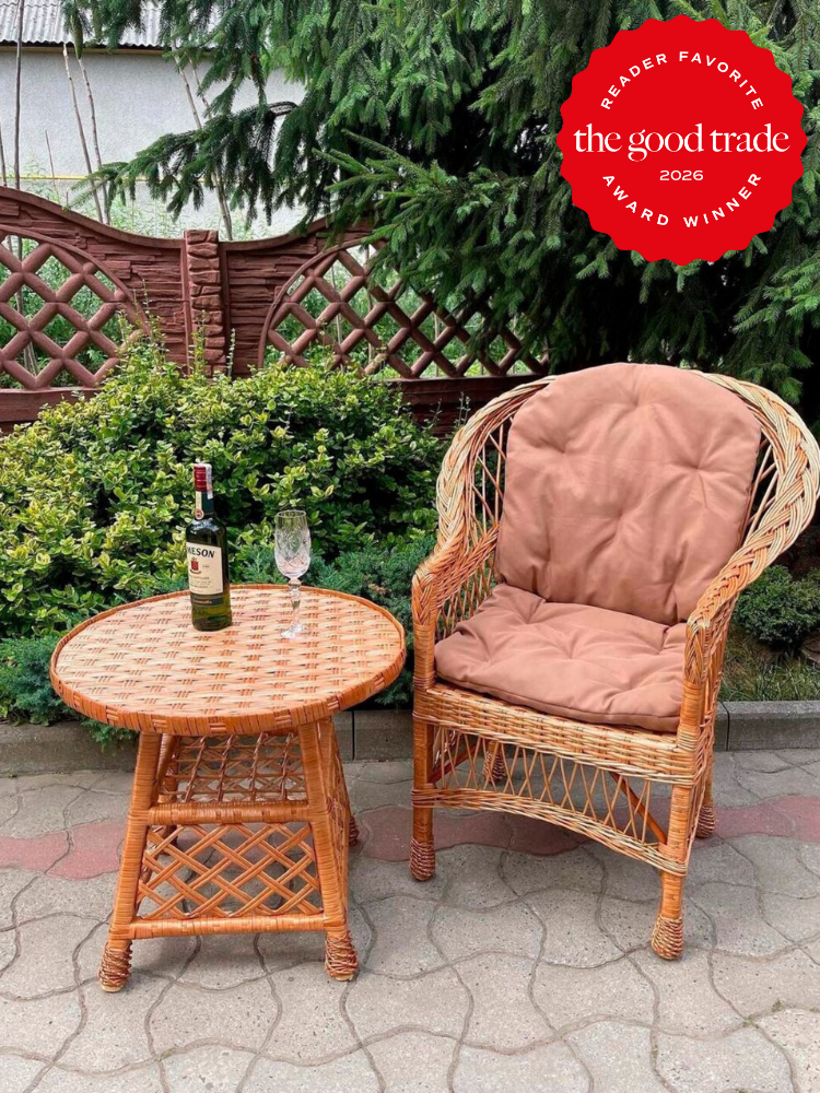 A wicker chair with a brown cushion and a round wicker table holding a wine bottle and glass, set on a patio with greenery and lattice fencing in the background. A red award badge appears in the corner.