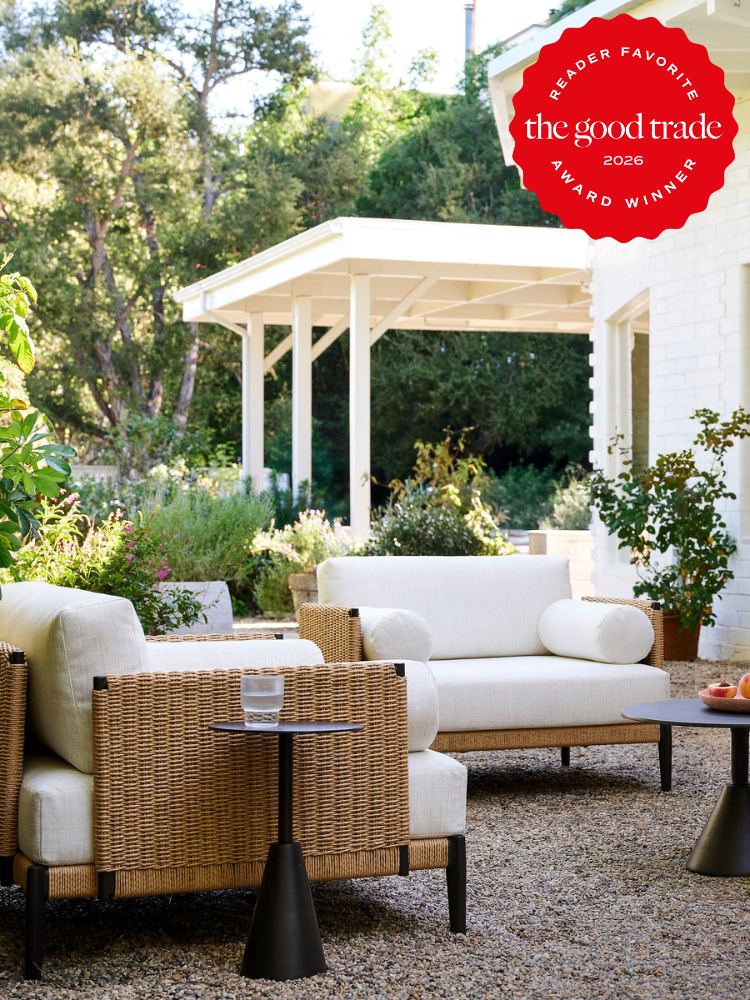Outdoor patio with wicker furniture and white cushions, small tables, gravel ground, garden plants, and a white building; "The Good Trade 2026 Award Winner" badge in the top right corner.