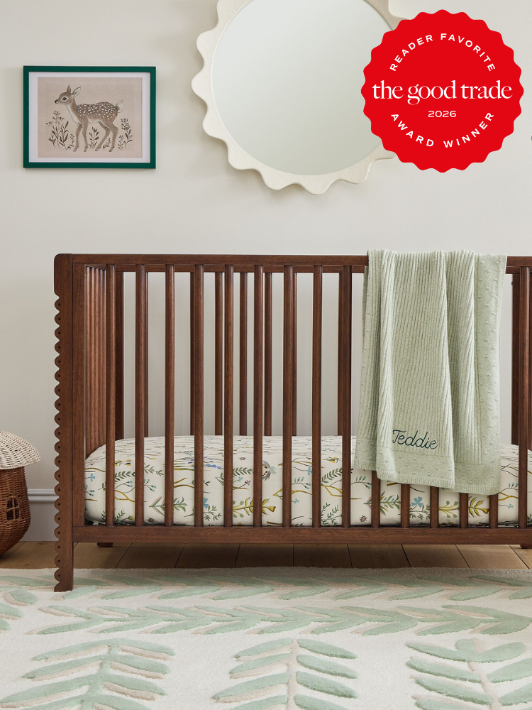 A wooden crib with a personalized green blanket and leaf-patterned bedding stands in a nursery with a deer picture, round mirror, and a green leaf rug. "The Good Trade 2026 Award Winner" badge.