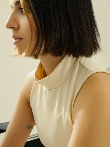 Person with short black hair and a blond streak, wearing a sleeveless beige top, looking sideways.