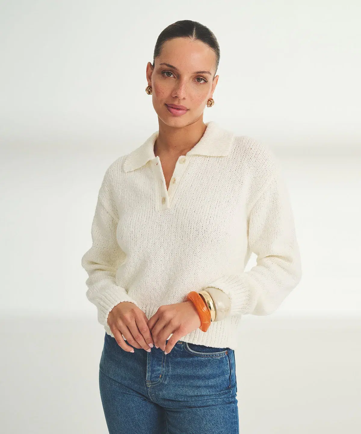 Woman wearing a cream knit sweater and blue jeans, standing against a plain white background, with hair pulled back and accessorized with earrings and bracelets.
