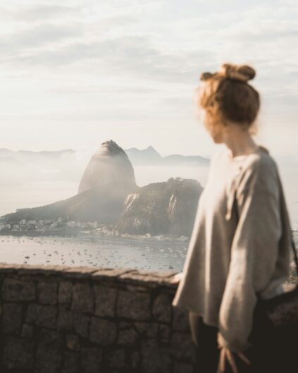 A person with light brown hair in a bun stands by a stone wall, overlooking Sugarloaf Mountain and the bay in Rio de Janeiro, Brazil, on a hazy day.