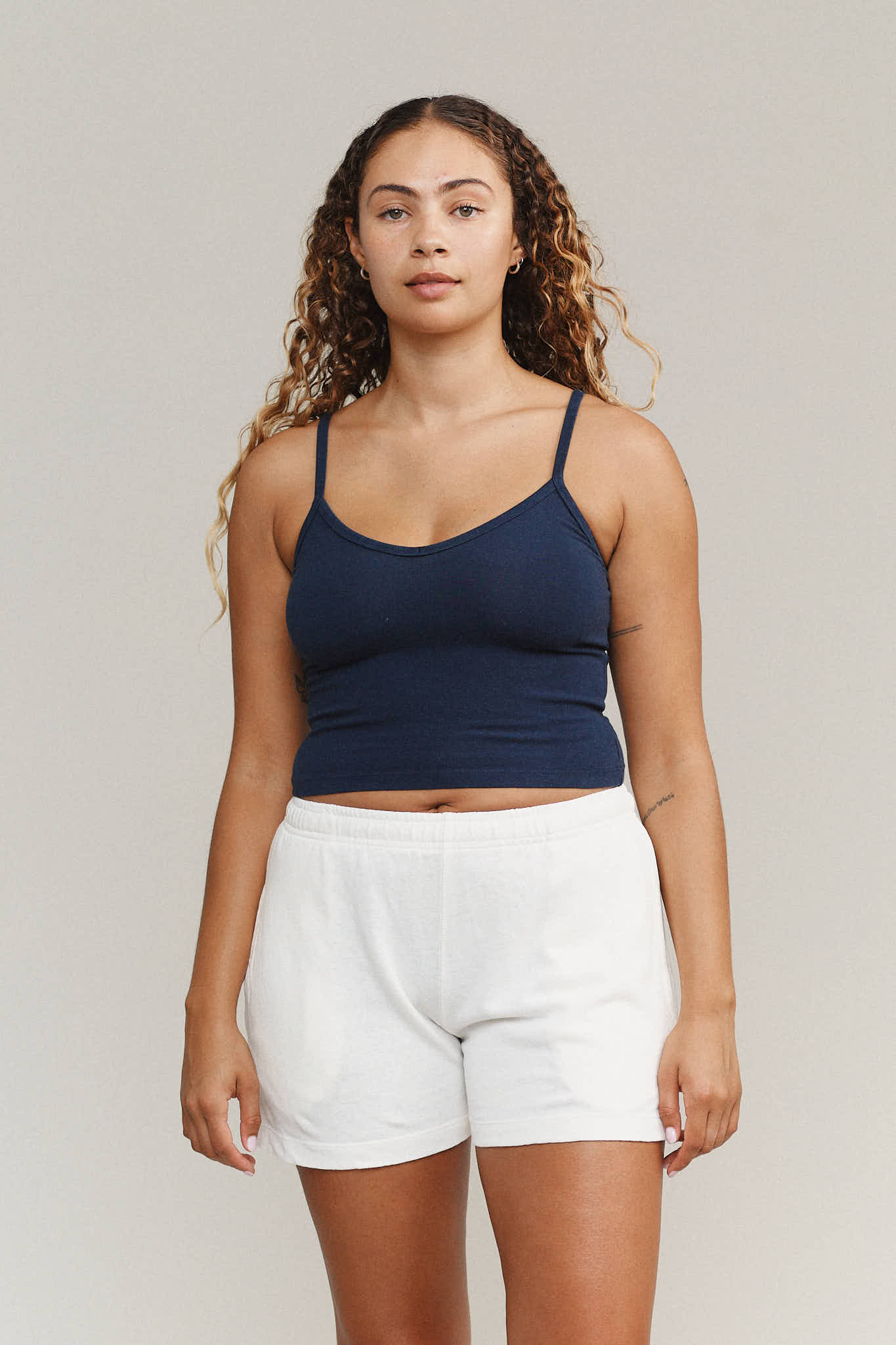 A person with long curly hair wearing a navy tank top and white shorts stands against a plain background.