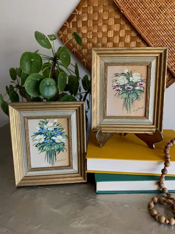 Two small framed floral paintings on a table, accompanied by stacked books, a beaded necklace, and a potted plant.