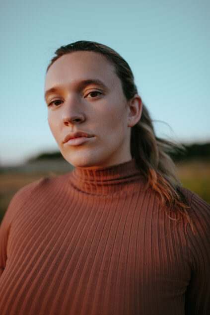 A person with long hair pulled back, wearing a ribbed brown turtleneck, stands outdoors during sunset with a neutral expression.
