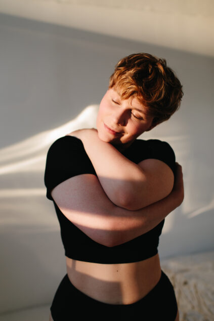 Person with short hair hugging themselves, standing against a white wall, wearing a black top, and bathed in warm light. Eyes are closed and a soft expression is on their face.