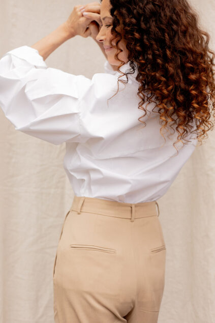 Person with curly hair wears a white blouse and beige pants, standing against a neutral backdrop.