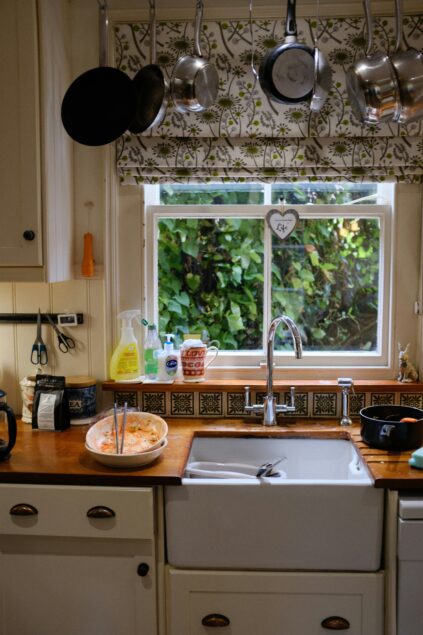 A kitchen sink with a wooden countertop, assorted dishes, cleaning supplies, and kitchen utensils. Pots and pans hang above, and a window overlooks leafy greenery outside.