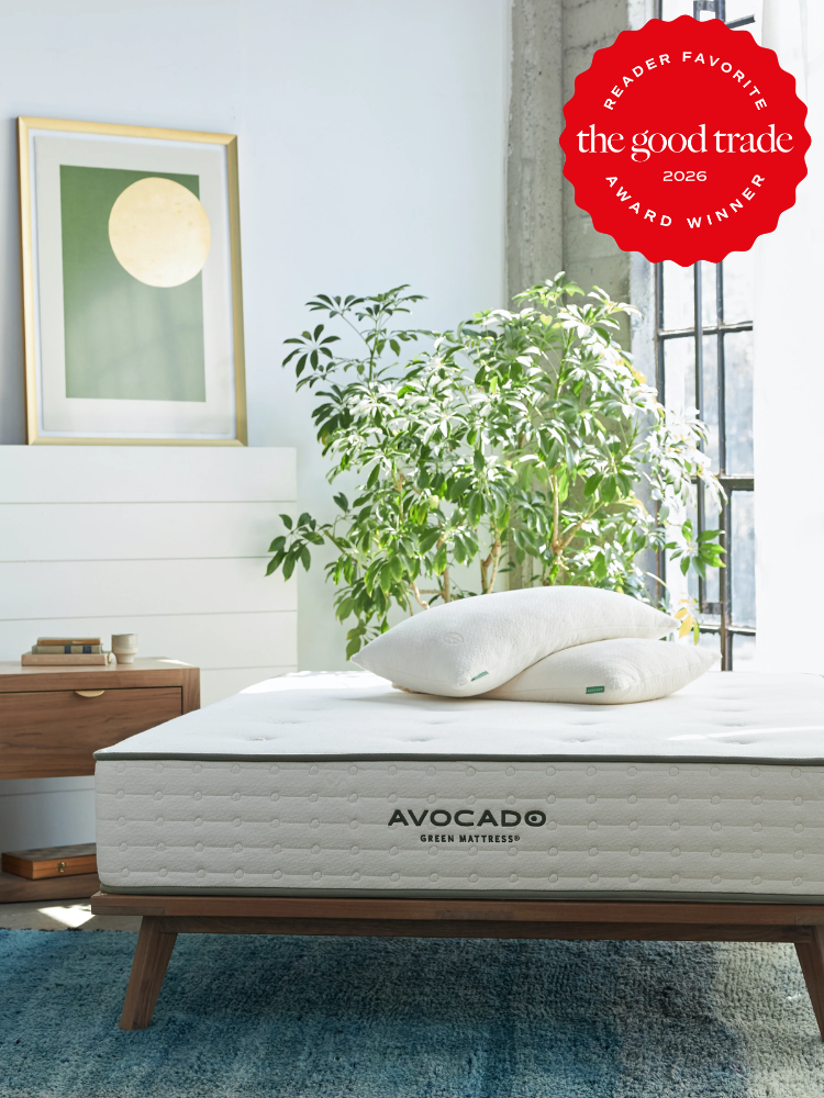 A neatly made Avocado Green Mattress with pillows on a wooden bed frame, in a bright room with a plant, nightstand, and framed art; award badge in the top right corner.