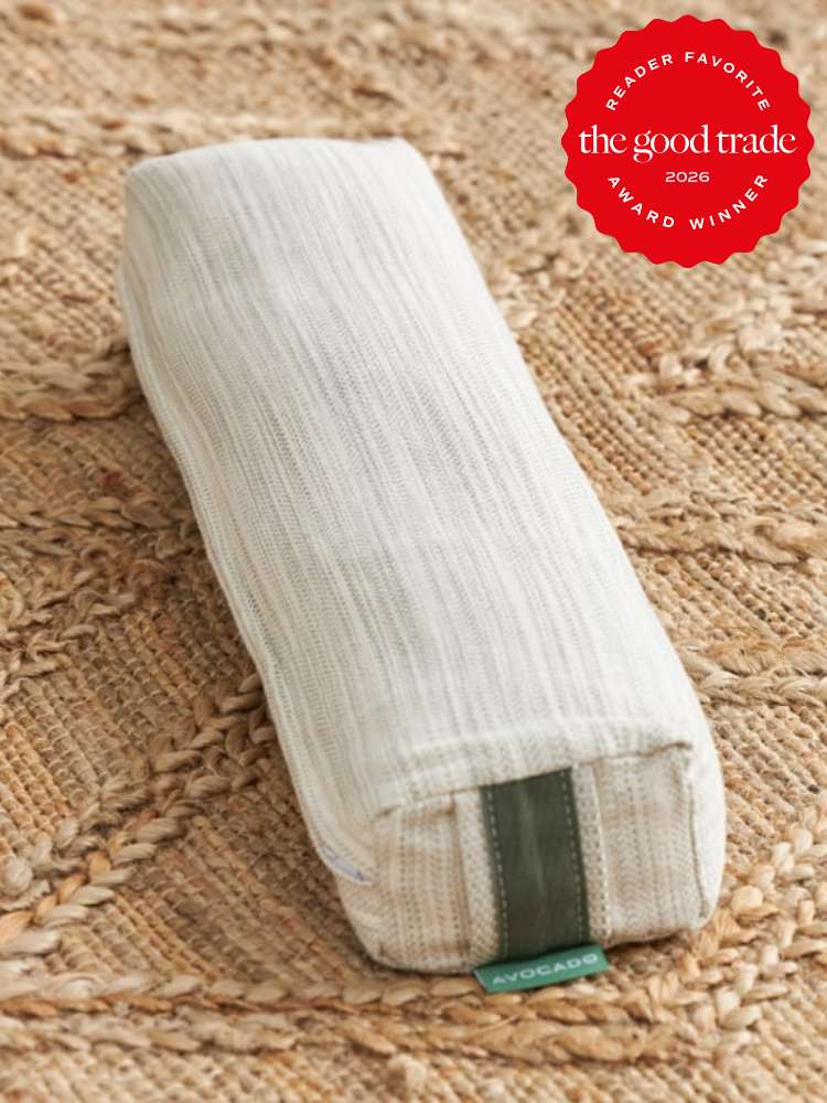 A rectangular, off-white fabric pillow with a green tag labeled "Avocado" rests on a woven rug. A red badge in the corner reads "Reader Favorite The Good Trade 2026 Award Winner.