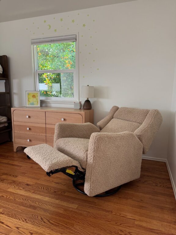 A beige recliner chair with an extended footrest sits on a wooden floor near a dresser and window in a softly lit room.