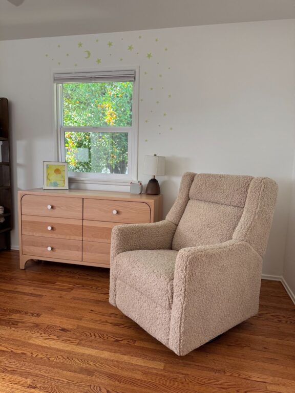 A beige upholstered armchair sits on a wooden floor next to a light wood dresser with a window and star decals on the white wall behind them.