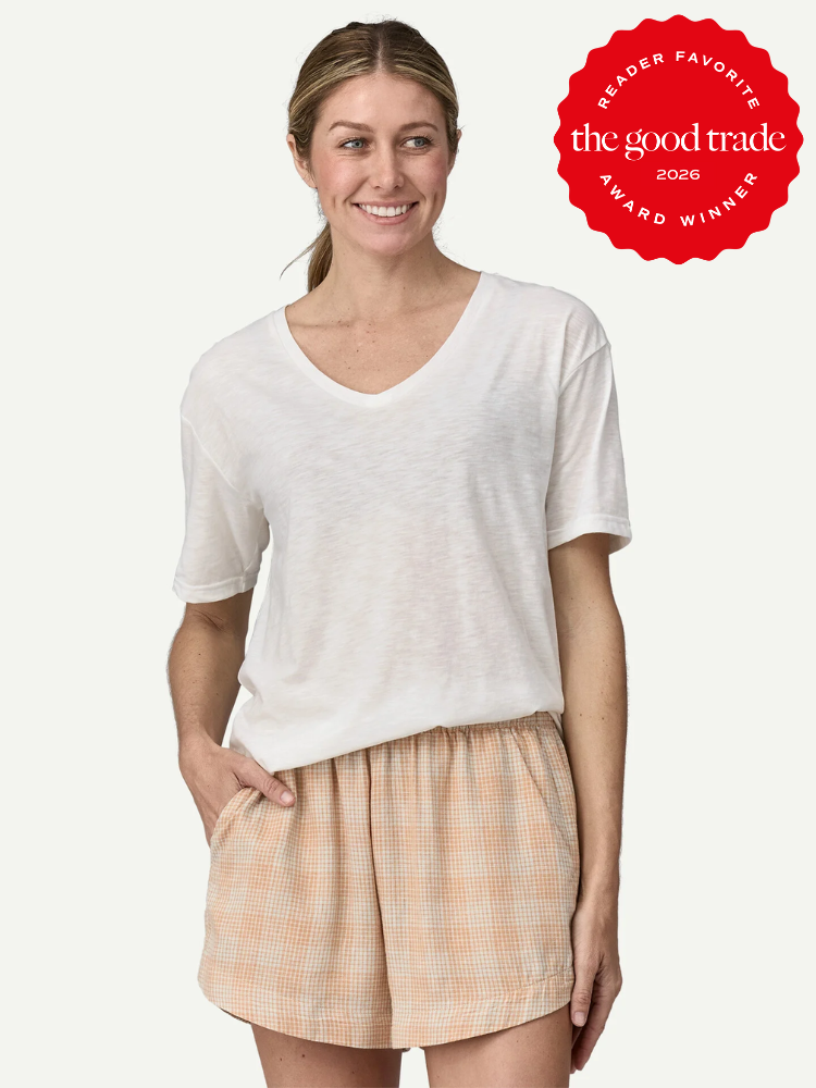 Woman wearing a loose white V-neck T-shirt and peach plaid shorts, standing with one hand in her pocket. “The Good Trade Award Winner 2026” badge is displayed in the upper right corner.