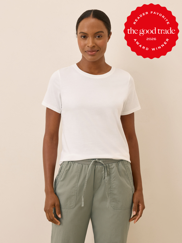 A woman stands against a plain wall, wearing a white t-shirt and sage green pants. A red "The Good Trade 2026 Award Winner" badge appears in the top right corner.