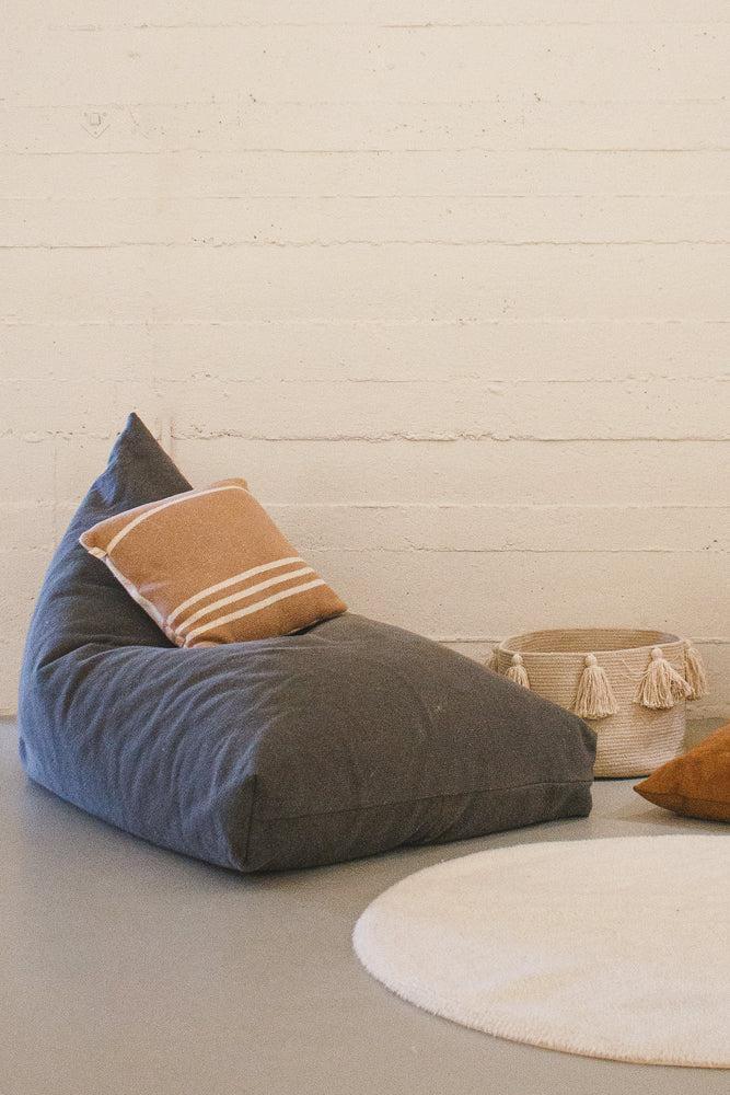 A gray bean bag with a brown striped cushion is placed on a light floor next to a round white rug and a decorative basket with tassels.