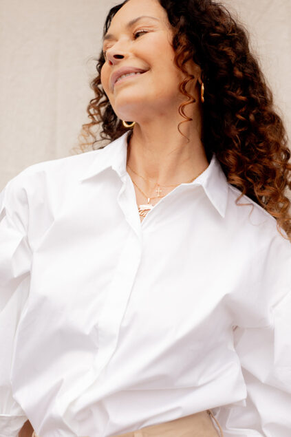 Woman with curly hair wearing a white button-down shirt and gold jewelry, smiling and looking to the side against a neutral background.