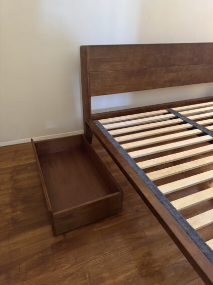 Wooden bed frame with horizontal slats and an extended storage drawer on a wooden floor.