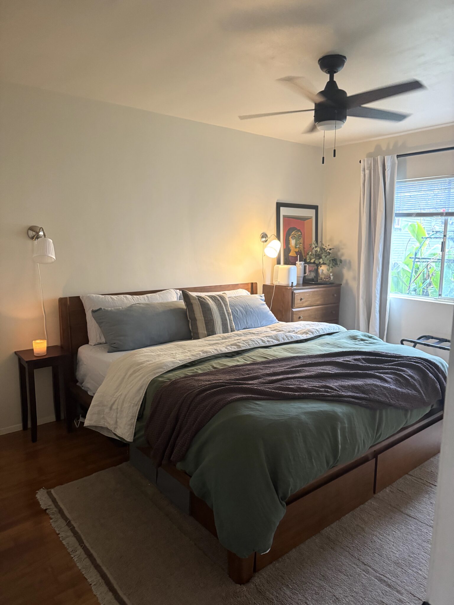 A neatly made bed with green and white bedding in a cozy bedroom, featuring a ceiling fan, nightstands, a dresser, framed artwork, and a window with natural light.