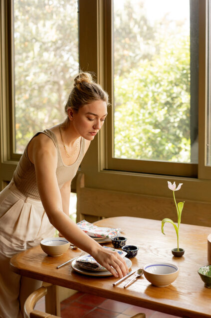 A woman arranges tableware on a wooden dining table set with bowls, plates, and chopsticks next to a window with greenery outside.