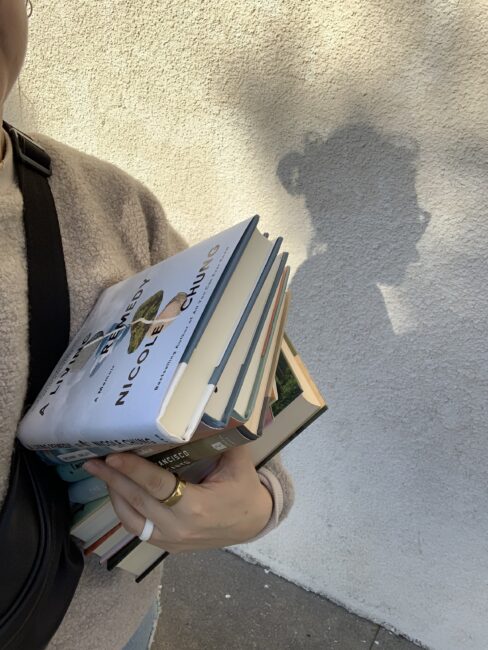 A person holding a stack of books, with “A Living Remedy” by Nicole Chung on top, against a textured wall.
