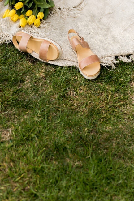A pair of tan sandals on a beige blanket next to a bouquet of yellow tulips, set on green grass.