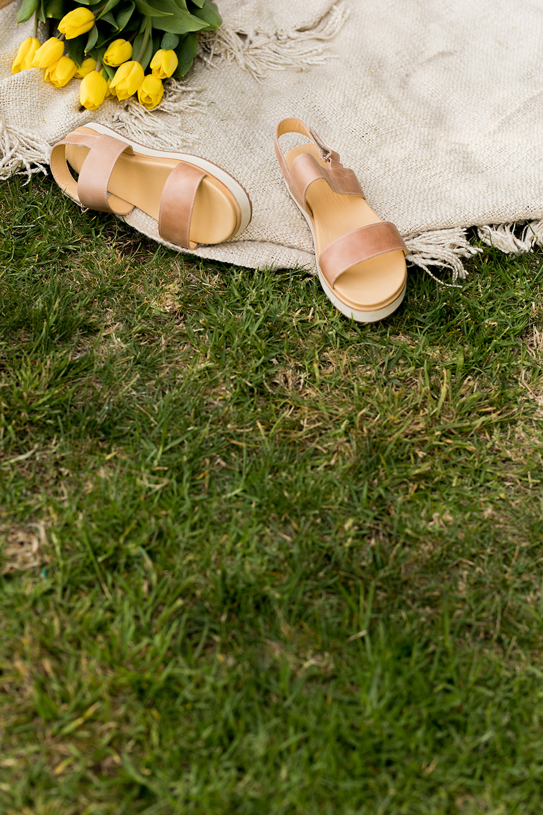 A pair of tan sandals on a beige blanket next to a bouquet of yellow tulips, set on green grass.
