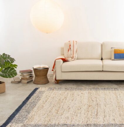 A beige sofa with a colorful throw and cushion sits against a white wall, beside a stack of books, a round lamp, a potted plant, and a woven basket on a rug.