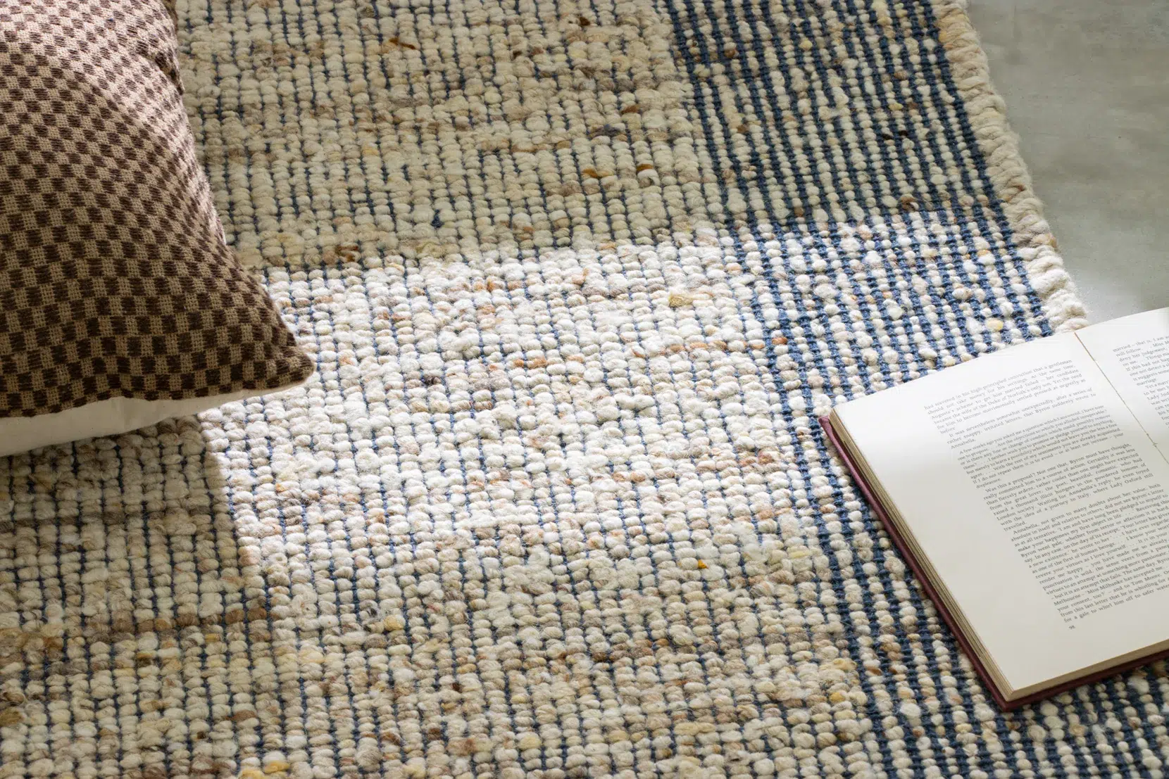 A checkered cushion, an open book, and a woven rug with blue and beige patterns are arranged on the floor with sunlight highlighting part of the rug.