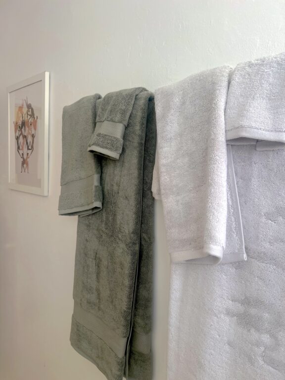 Two organic towels, one gray and one white, hang side by side on a rack against a white wall; a framed artwork is visible on the left side of the image.