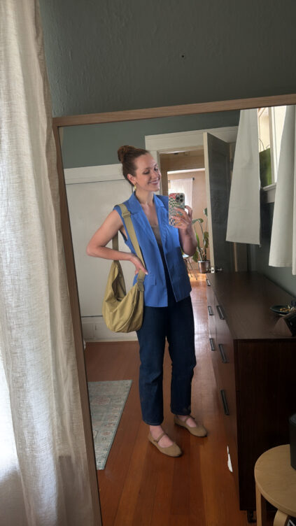 Woman stands in front of a mirror, smiling and taking a selfie. She is wearing a blue sleeveless top, dark pants, beige flats, and carries a tan shoulder bag.