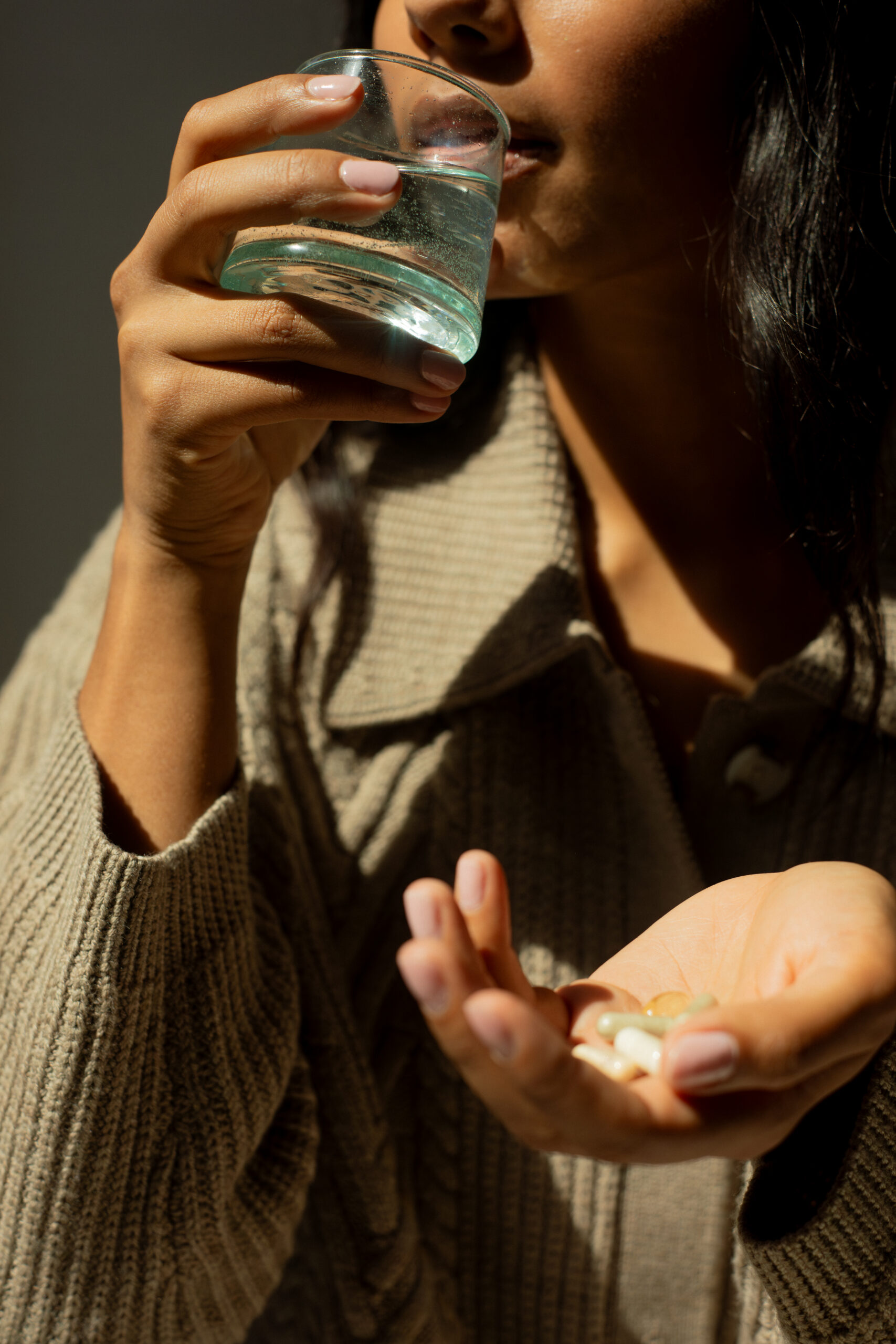 A person holds several capsules in one hand and lifts a glass of water to their mouth with the other hand, wearing a beige sweater.