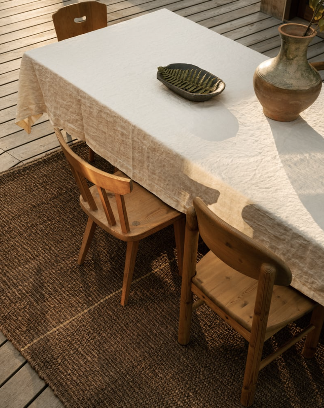 Wooden dining table with a white tablecloth, ceramic vase, and leaf-shaped dish, surrounded by wooden chairs on a textured brown rug.