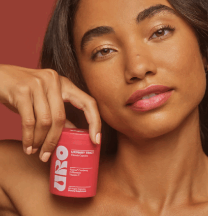 A woman holds a pink container of urinary tract vitamin capsules labeled "URO" close to her face against a solid reddish background.