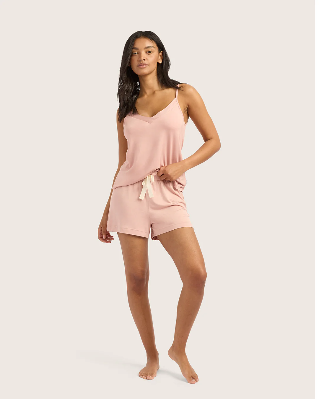 A woman stands barefoot wearing a light pink camisole and matching shorts with a drawstring, posed against a plain background.