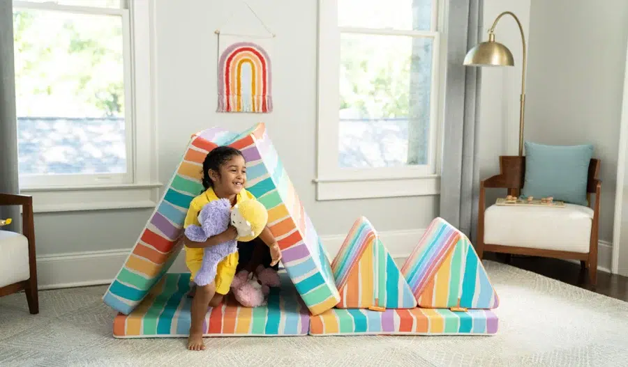 A child holding stuffed animals stands in a colorful, striped play tent in a bright living room with large windows and simple furniture.