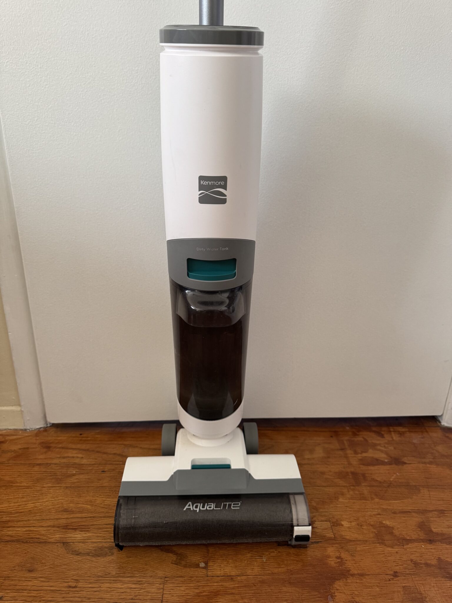 A white and gray Kenmore AquaLite upright vacuum cleaner stands on a wooden floor in front of a white wall.