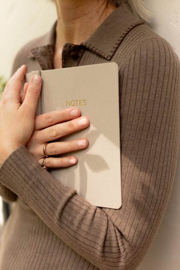 A person wearing a ribbed brown sweater holds a beige notebook labeled "NOTES" against their chest with both hands.