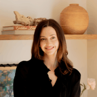 A woman with long brown hair and a black top smiles in front of a wooden shelf with books, a clay pot, driftwood, and a framed picture.