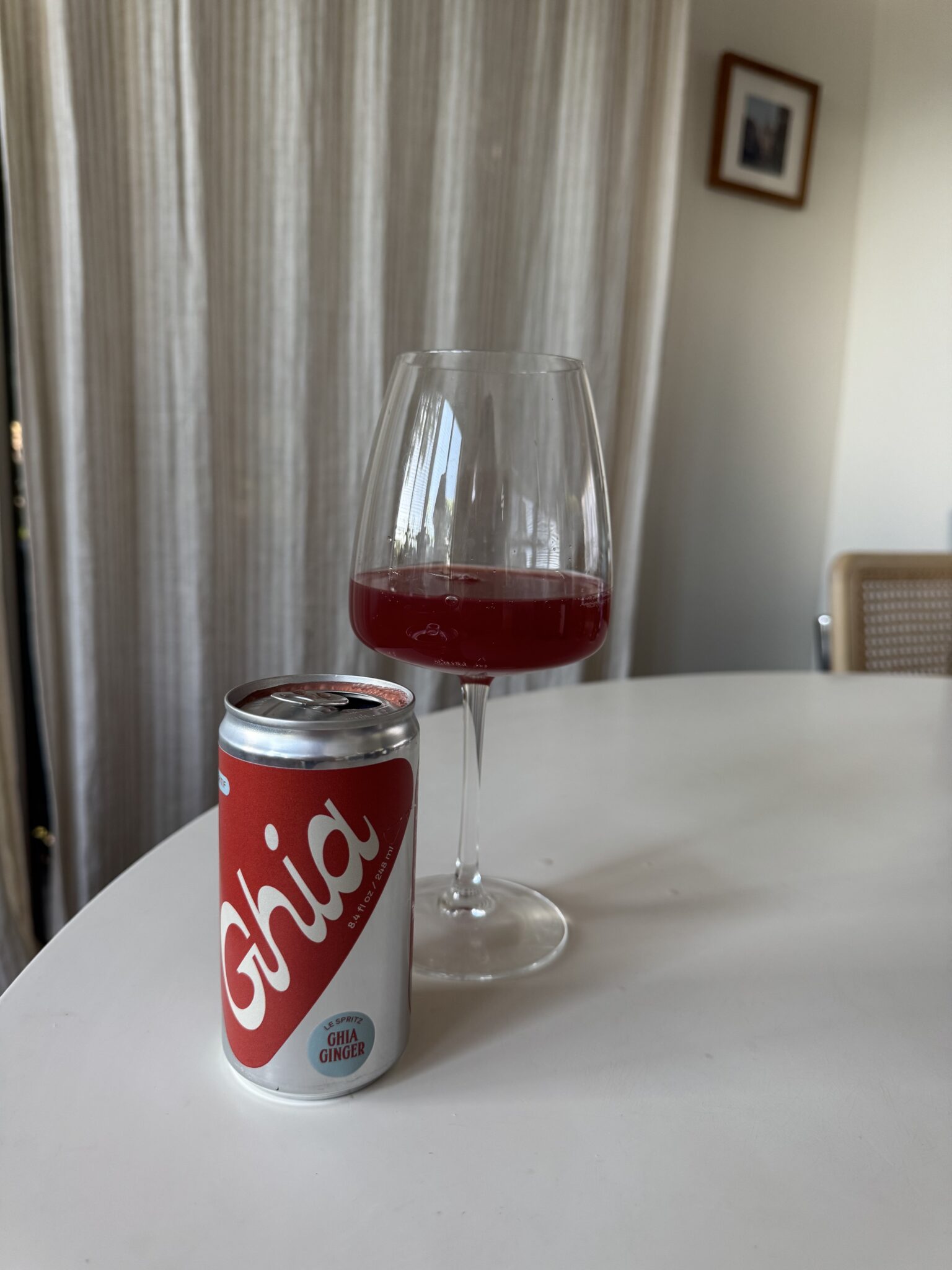 A can of Ghia ginger soda and a wine glass filled with a red beverage sit on a white table in a room with light curtains and a framed picture on the wall.