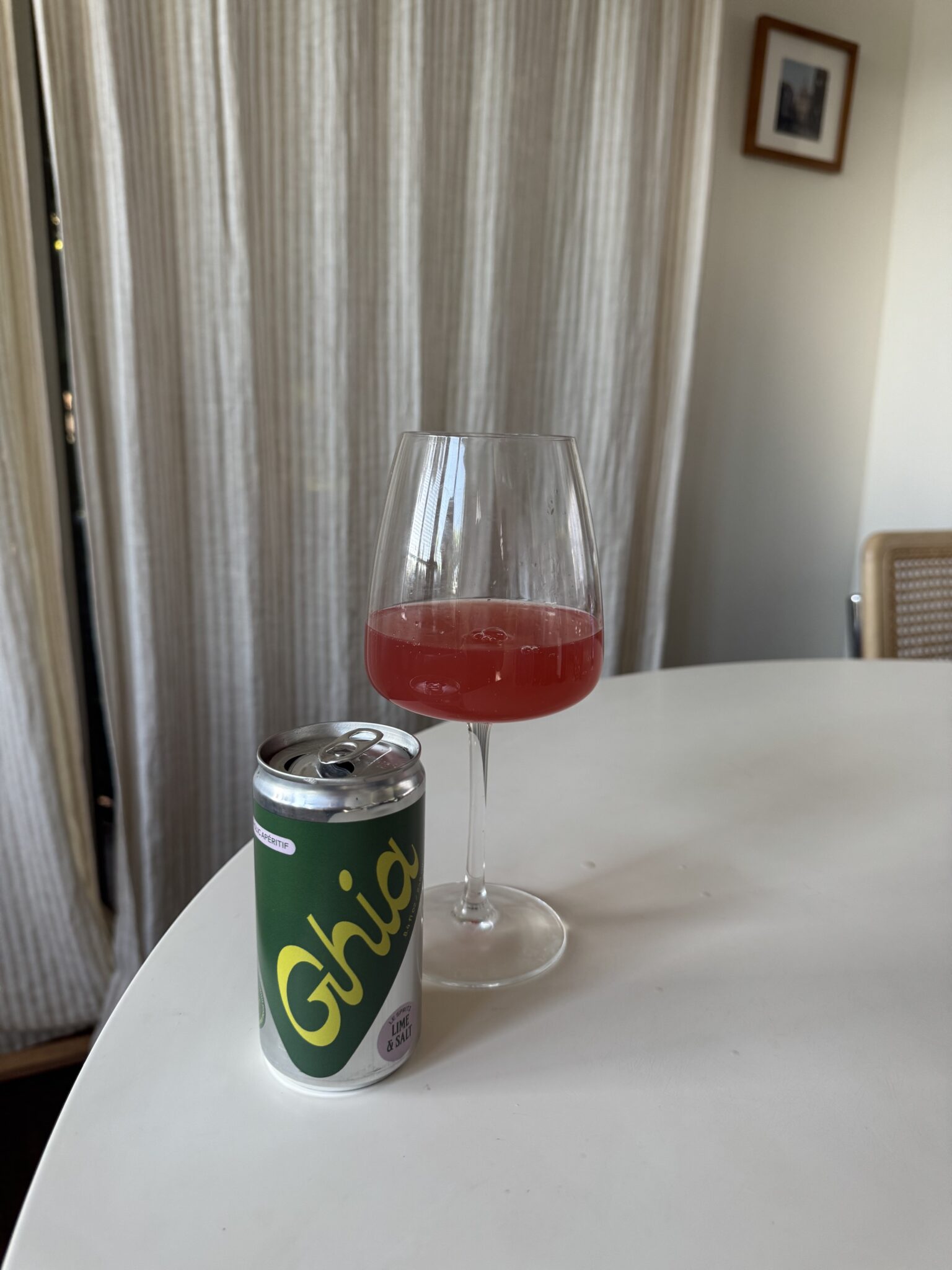A can of Ghia soda and a wine glass with a red beverage sit on a white table in a softly lit room.