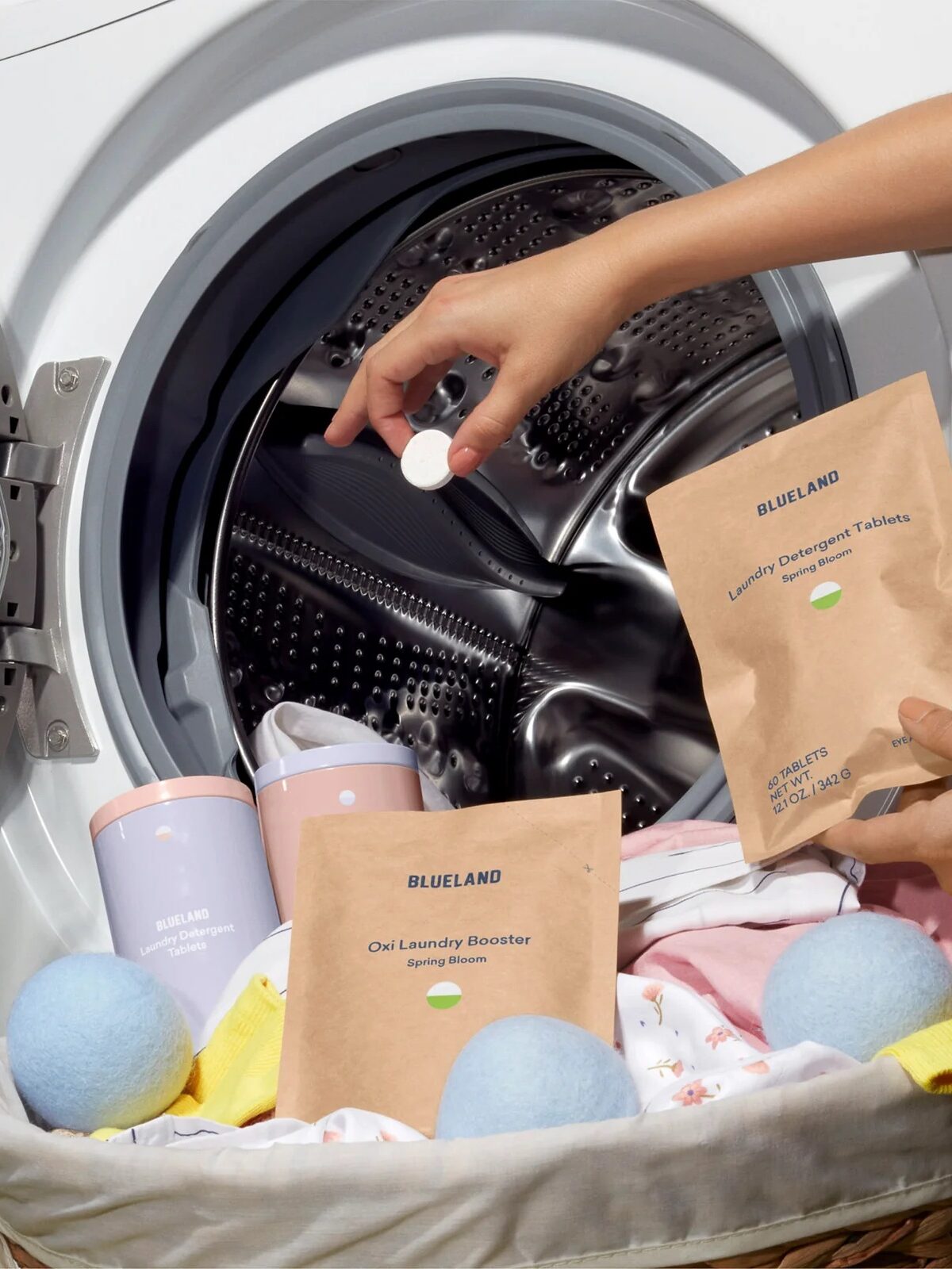 A person places a laundry tablet into a front-load washing machine with laundry inside. Laundry products and wool dryer balls are also visible.