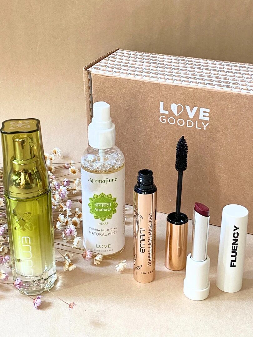 A set of beauty products including a serum, mist, mascara, and lipstick is displayed in front of a LOVE GOODLY box on a beige surface with small dried flowers.