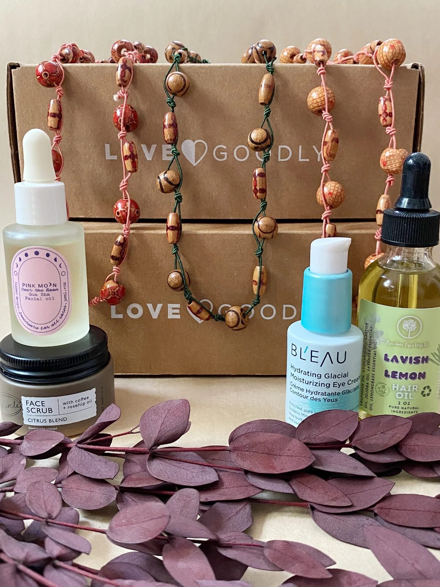 A selection of skincare products, two beaded necklaces, and dried leaves displayed in front of two stacked Love Goodly boxes.