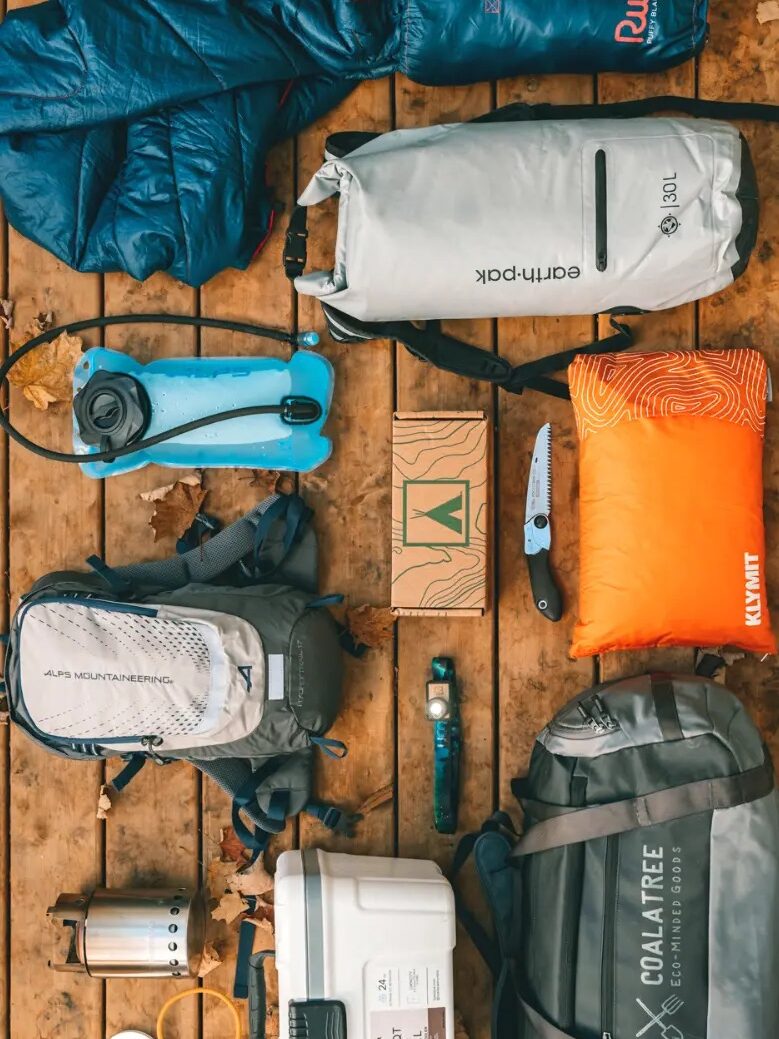 Various camping gear including a cooler, backpack, sleeping bag, water reservoir, duffel bag, lantern, flashlight, knife, and pillow arranged on wooden decking.