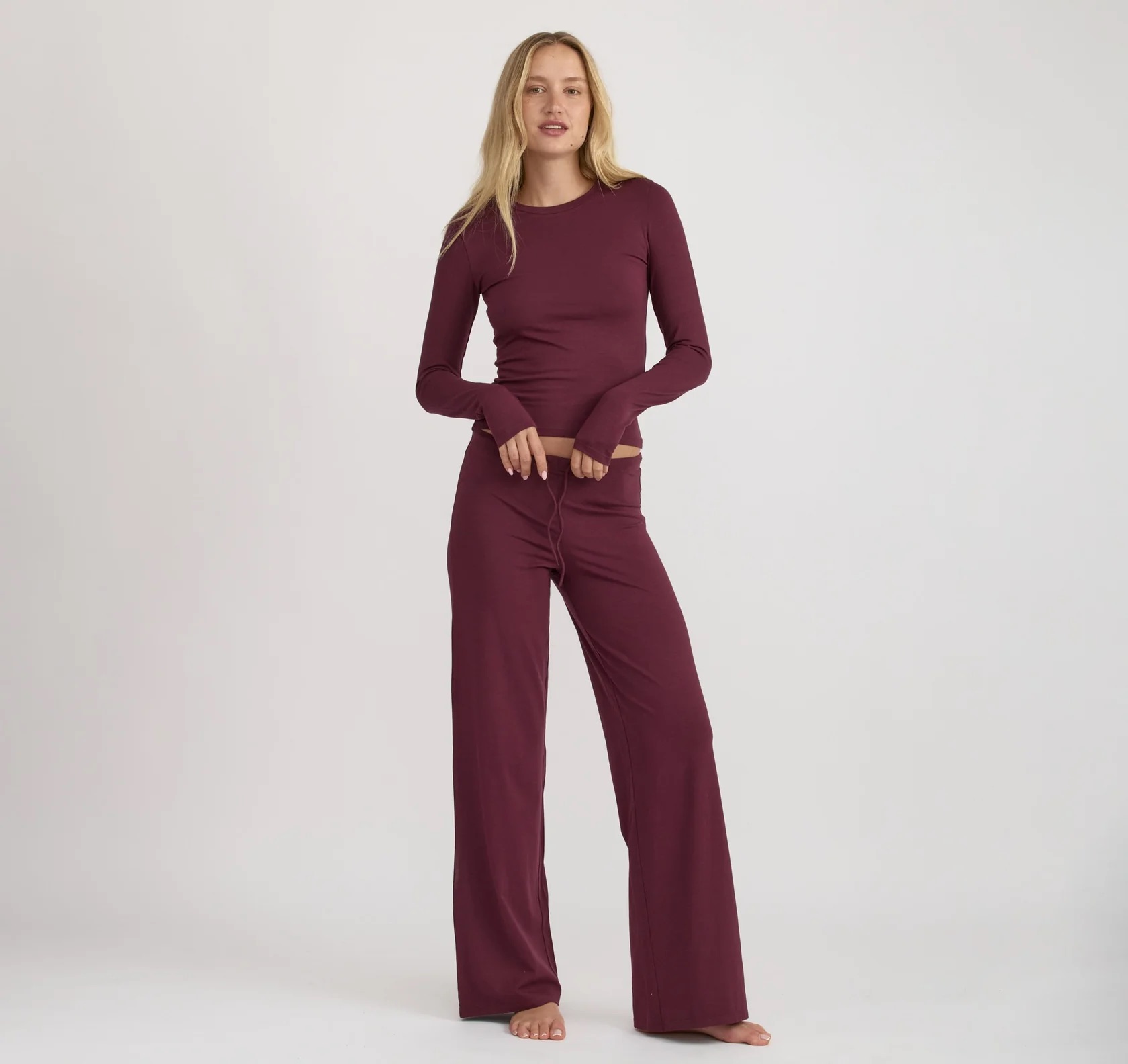 A woman stands barefoot against a plain background, wearing a long-sleeve, burgundy top and matching wide-leg pants.